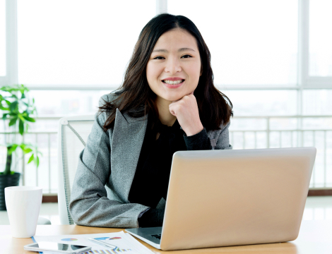 A woman in professional attire seated at a desk, working on a laptop.